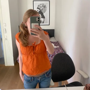 Orange linnetopp från Debbie Katz - Superfin orange linnetopp från Debbie Katz med korta ärmar och söt knytning framtill. Toppen har en luftig och lätt struktur, perfekt för varma dagar. Den har en avslappnad passform och är tillverkad i mjukt linne som känns skönt mot huden.