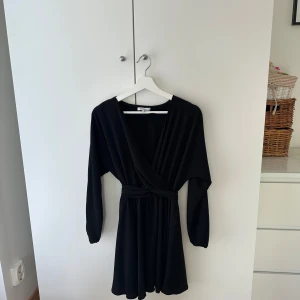 Svart omlottklänning från Chiquelle - Svart kort omlottklänning från Chiquelle med långa ärmar och knytband i midjan. Klänningen har v-ringning och en flowy kjol som ger en snygg siluett. Perfekt för dig som gillar stilrena och enkla plagg med en twist.