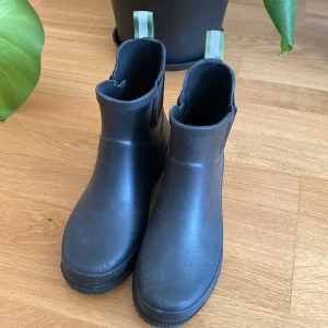 Svarta gummistövlar från Roots - Svarta gummistövlar från Roots med klassisk rund tå och elastiska paneler på sidorna. Praktiska gröna och gråa dragflikar baktill gör dem enkla att ta på. Perfekta för regniga dagar och snygga till alla outfits. Något dammiga och använda av mig utomhus ca 2 ggr.