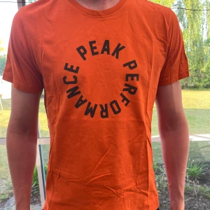 Orange Peak Performance t-shirt - Snygg orange t-shirt från Peak Performance med svart cirkelformad text på bröstet. Klassisk rund halsringning och korta ärmar. Skön passform och tillverkad i mjuk bomull, perfekt för en avslappnad stil.