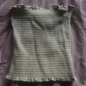 Beige bandeau tubtopp från H&M - Supersöt beige bandeau tubtopp från H&M i smockad bomull med volangdetaljer upptill och nedtill. Perfekt att styla med höga jeans eller kjol för en chill och trendig look. Toppen är ärmlös och har en tight passform som sitter snyggt.