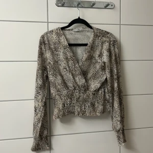 Beige omlottblus med djurmönster - Trendig beige omlottblus med svart och brunprickigt djurmönster. Blusen har v-ringning, långa ärmar och smockad midja som ger en snygg passform. Perfekt för dig som gillar en cool och avslappnad stil.