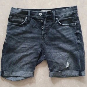 Jeansshorts - Snygga grå svart jeansshorts från H&M med klassisk femficksdesign, uppvikta benslut och en liten sliten detalj framtill. Shortsen har normal passform och stängs med knappar. Perfekta för en chill sommardag.