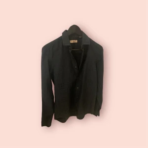 Svart skjorta från Burberry London - Stilren svart skjorta från Burberry London med klassisk krage och knäppning framtill. Skjortan har lång ärm och är tillverkad i ett mjukt bomullstyg. Perfekt för dig som gillar en clean och tidlös look. Storlek 15 - 38 vilket motsvarar M. Armhåla till armhåla 44cm. Totallängd 68