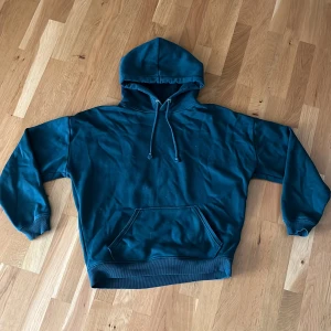Mörkgrön hoodie från BikBok - En mörkgrön hoodie från BikBok i storlek M. Tröjan har huva med snörning, magficka och ribbade muddar. Tillverkad i mjuk bomullsmix som känns skön mot huden. Perfekt för en chill och avslappnad stil. (Ljuset i bilden får hoodien att se mörkblå ut)