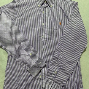 Randig skjorta Polo Ralph Lauren M - Snygg lila och vit randig skjorta från Polo Ralph Lauren i slim fit. Klassisk button-down krage, långa ärmar och broderad logga på bröstet. Skjortan är i bomull och har knappar framtill. Perfekt för dig som gillar stilrena och fräscha plagg.