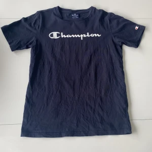 Mörkblå Champion t-shirt i bomull - Snygg mörkblå t-shirt från Champion med stort vitt logotryck över bröstet och klassisk rund hals. T-shirten är i mjuk bomull och har korta ärmar med Champion-logga även på ärmen. Perfekt för en chill och sportig look.