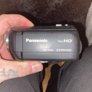 Videokamera Panasonic - Bilderna visar en Panasonic HC-V160 videokamera med Full HD, 8.9 megapixlar och 77x i.Zoom. Kameran har utfällbar skärm, HDMI- och AV-utgång samt enkel hantering. Perfekt för dig som vill skapa egna filmer eller vloggar med bra bildkvalitet.