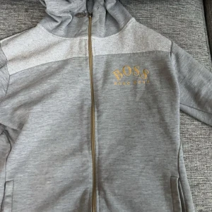 Grå zip-hoodie från Hugo Boss S - Snygg grå hoodie från Hugo Boss med guldfärgad dragkedja och logga på bröstet. Jackan har huva, fickor och är tillverkad i mjuk bomullsblandning. Perfekt för en chill och stilren look. Fråga i dm om mer bilder plus finns att de står från Hugo boss där vid storleken 