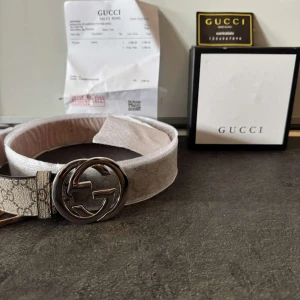 Beige GG-mönstrat bälte från Gucci - Snyggt beige bälte från Gucci med klassiskt GG-mönster och stor silverfärgad logga som spänne. Bältet är gjort i canvas med detaljer i skinn och har en stilren, lyxig känsla. Perfekt accessoar för att lyfta din outfit med en exklusiv touch.