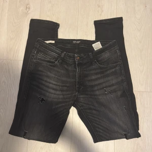 Svarta skinny jeans från Jack & Jones - Snygga svarta skinny jeans från Jack & Jones med slitningar på benen för en cool look. Jeansen har klassisk femficksdesign, knappgylf och är tillverkade i stretchigt denim för extra komfort. Perfekta för dig som gillar en smal passform och streetstil. Storlek 34/34 sitter som 31/32