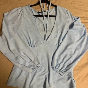 Ljusblå långärmad blus med knyt - Supersöt ljusblå blus med v-ringning och långa puffärmar. Toppen har knytband i nacken och lätt strukturerat tyg som ger en extra touch. Perfekt för dig som gillar en feminin och trendig stil.