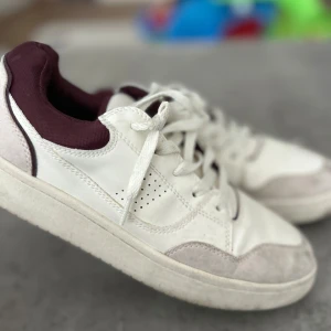 Vita sneakers med vinröda detaljer - Snygga vita sneakers med vinröda detaljer runt ankeln och perforerade sidor för extra ventilation. Skorna har snörning, rund tå och platt sula. Ovandelen är i syntet och textil, med förstärkningar i mocka framtill och baktill. Perfekta för en stilren och sportig look.