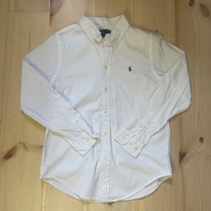 Ralph Lauren Skjorta - Klassisk vit skjorta från Ralph Lauren med button down-krage och broderad marinblå logga på bröstet. Skjortan har lång ärm, knappar framtill och är tillverkad i bomull. Perfekt för en clean och stilren look.