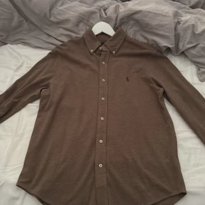 Brun skjorta från Polo Ralph Lauren - Snygg brun skjorta från Polo Ralph Lauren i lätt och mjuk bomull. Klassisk button-down krage, långa ärmar och knäppning framtill. Diskret broderad logga på bröstet. Perfekt för dig som gillar stilrena och tidlösa plagg. Aldrig använt skulle säga att den passar bra på fina middagar! 