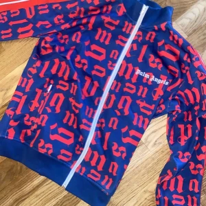 Palm Angels blå och röd track tracksuit - Palm Angels track jacket i blått med rött grafiskt mönster och vita detaljer. Jackan har hel dragkedja framtill, ståkrage och röda ränder längs ärmarna. Tillverkad i bomull och polyester för en sportig vibe. Perfekt för dig som gillar streetwear med attityd. ALDRIG använt passar inte mig de står s fast passar mellan s och m jätte fina och billiga.