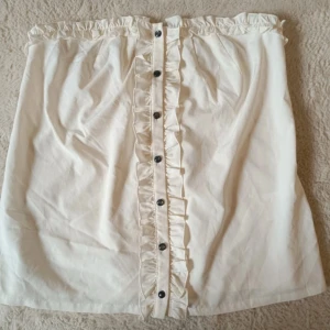 Vit blus med volang och knappar - Supersöt vit tube top ( blus )  med volangdetalj längs knappraden framtill. Blusen har en rak passform och är perfekt för dig som gillar en feminin och trendig stil. Lätt att matcha med både sneakers och boots. Priset är diskuterbart! 