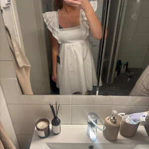 Vit kortklänning med volang från Lulus - Supersöt vit kortklänning från Lulus med volangdetaljer på axlarna och spetskant vid urringningen. Klänningen har en luftig passform, fyrkantig ringning och knytning i ryggen. Perfekt för sommarens alla tillfällen!