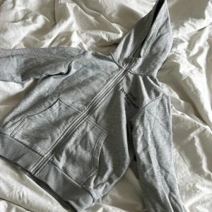 Grå hoodie från Peak Performance - Snygg grå hoodie från Peak Performance med dragkedja och broderad logga på bröstet. Tröjan har huva, långa ärmar och två stora fickor framtill. Perfekt för chill dagar eller när du vill ha en sportig look.