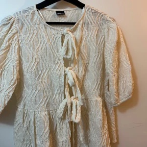 Vit söt tröja/topp från Gina Tricot - Supersöt vit kortklänning från Gina Tricot med strukturmönster och tre knyt framtill. Tröjan har voluminösa ärmar och en luftig känsla, perfekt för varmare dagar. Materialet är lätt och ger en cool textur till outfiten. Tog bort etiketten men aldrig använt. Storlek S. köpte den för 400kr