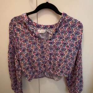 Blommig omlottblus från Zara - Säljer en blommig omlottblus från H&M med långärmade puffiga ärmar och croppad passform. Blusen har ett färgglatt mönster i blått, rosa och vitt, och är gjord i tunn bomull. Perfekt för dig som gillar boho-vibe och snygga detaljer.
