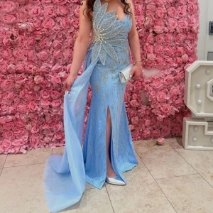 Ljusblå glittrig långklänning med slits - Säljer en ljusblå långklänning med glittriga detaljer och pärlor framtill. Klänningen har smala axelband, en v-ringad omlott-topp och en elegant slits framtill. Materialet är glittrigt och lätt, med ett långt släp som ger extra wow-effekt. Använd endast en gång! Priset kan diskuteras. Nypris 6000!