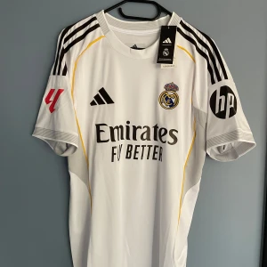 Real Madrid Arda Güler matchtröja 25/26 - Säljer en Real Madrid fotbollströja från säsongen 25/26, med Arda Güler och nummer 15 på ryggen. Tröjan är vit med svarta detaljer, gula linjer och tryck på ärmarna. Slim fit-modell i lätt, ventilerande polyester. Klassisk rund hals och korta ärmar.