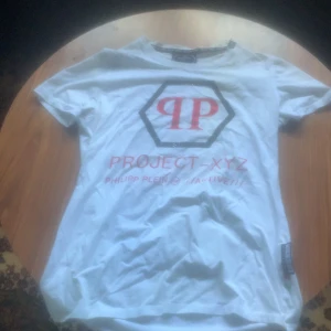 Vit Philipp Plein Project-XYZ t-shirt - Snygg vit t-shirt från Philipp Plein med Project-XYZ tryck och PP-logga i rött framtill. Baksidan har cool döskalle och streckkod i rött. Klassisk passform och korta ärmar, perfekt för dig som gillar streetstyle och statement-plagg.