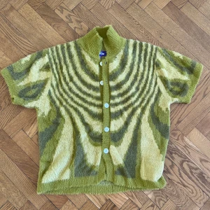 Grön mönstrad kofta från Fucking Awesome - Säljer en unik och fluffig kofta från Fucking Awesome i storlek XS. Koftan har ett grönt och beige psykedeliskt mönster, korta ärmar och vita knappar framtill. Materialet är stickat och har en mjuk, lurvig känsla.