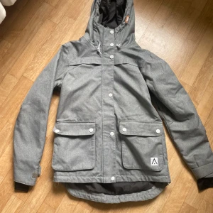 Grå vindjacka från WearColour - Snygg grå vindjacka från WearColour med huva, stora fickor med lock och både dragkedja och knappar framtill. Jackan har en loose passform och är tillverkad i slitstark polyester, perfekt för dig som gillar streetstyle och funktion. Fodrad insida för extra komfort.