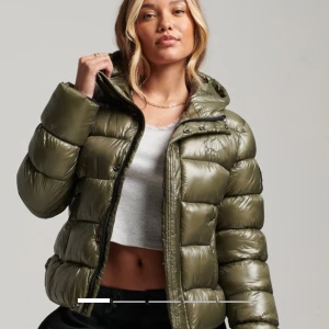 Olivgrön pufferjacka från superdry  - Snygg olivgrön pufferjacka från superdry med huva och dragkedja framtill. Jackan har quiltad design, två fickor och ribbade muddar vid ärmslut. Perfekt för kalla dagar och har en cool, lite glansig finish som sticker ut. Storlek 42 men passar också en s