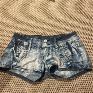 Blå jeansshorts - Snygga blå jeansshorts från Vero Moda med coolt tvättad effekt och silverfärgade knappar. Shortsen har låg midja, fram- och bakfickor med dekorativa detaljer och bälteshällor. Perfekta för varma dagar och en avslappnad stil.