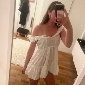 Vit volangklänning med knappar - Söt vit klänning med volangdetaljer vid axelbanden och knappar framtill. Klänningen är ärmlös och har en luftig, lätt form med broderade detaljer och en kort längd. Perfekt för varma dagar och somriga tillfällen.