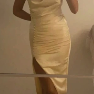 Gul festklänning  - Säljer en elegant klänning i gulfärgad satin med snygg drapering och dekorativa knappar längs sidan. Klänningen har en hög slits och är figurnära i passformen.