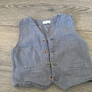 Grå väst från Newbie, stl 98 - Snygg grå väst från Newbie i bomull med diskret mönster och träknappar framtill. Västen har två fickor fram och dekorativa knappar bak. Perfekt för att ge outfiten en klassisk touch.