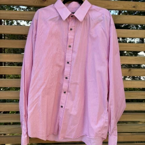 Rosa randig skjorta Riley XL - Snygg rosa skjorta från Riley i storlek XL med tunna vita ränder. Comfort fit-modell med klassisk krage och svarta knappar framtill. Perfekt för dig som gillar en lite loose passform.