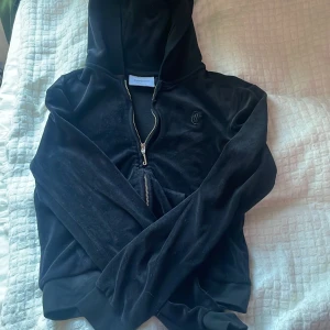 Svart velourjacka Juicy Couture - Svart hoodie från Juicy Couture med huva och hel dragkedja framtill. Klassisk JC-logga broderad på bröstet. Mjuk och skön känsla, perfekt för chill eller streetstyle. Ribbade muddar vid ärmslut och nederkant. Snygg och tidlös design. I storlek 10/11
