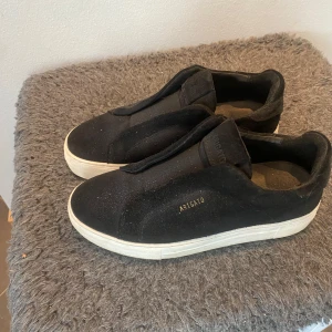 Svarta sneakers från Axel Arigato - Svarta sneakers från Axel Arigato med stilren design och vit platt sula. Skorna är gjorda i mocka och har en minimalistisk look utan synlig snörning. Perfekta för dig som gillar cleana och moderna sneakers med diskret branding i guld på sidan.