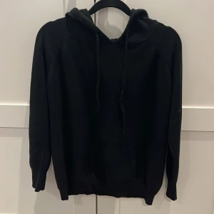 Svart merinoull hoodie - Svart merinoull hoodie med klassisk huva och snörning framtill. Tröjan har långärmade ärmar och ribbade muddar vid ärmslut och nederkant. Perfekt för chill dagar och enkel att matcha med jeans eller joggers. Materialet känns mjukt och skönt mot huden.