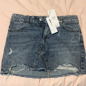 Blå jeanskjol från Gina Tricot - Helt ny blå jeanskjol, Från Gina Tricot med rå kant och slitna detaljer framtill. Kjolen har klassisk femficksdesign, knapp och dragkedja samt två bakfickor med lock och knapp. 