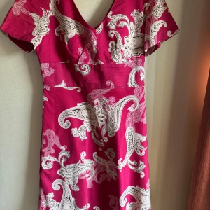 Röd mönstrad klänning Ann Taylor - Snygg röd klänning från Ann Taylor med vit paisley-inspirerat mönster. Klänningen har v-ringad hals, korta ärmar och figurnära passform. Tillverkad i mjuk bomull som känns skön mot huden. Perfekt för dig som gillar färg och mönster!