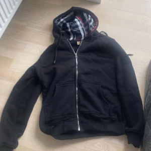 Svart hoodiejacka från Burberry - Svart hoodiejacka från Burberry med klassiskt rutigt foder i huvan i svart, vitt och rött. Jackan har dragkedja framtill, snörning i huvan och två stora fickor. Mjuk och skön bomullskänsla, perfekt för chill dagar. Ikonisk Burberry-logga broderad på bröstet.