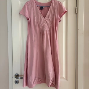 Rosa omlottklänning House of Lola - Söt rosa kortklänning från House of Lola med omlott-v-ringning och korta ärmar. Klänningen har rynkade detaljer längst ner och en liten broderad blomma vid fållen. Tillverkad i mjuk bomull, perfekt för vår och sommar.