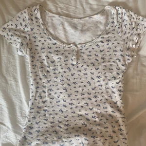 Blommig ribbad topp från brandy Melville  - Supersöt topp i ribbad bomull från brandy Melville , med små blå blommor på vit botten. Rund halsringning och tre knappar framtill🤍