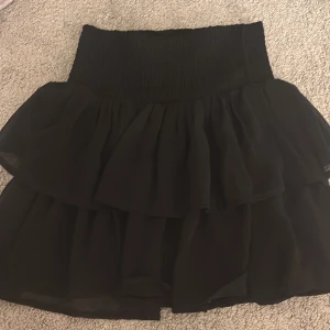 Svart volangkjol från KappAhl - Supersöt svart kjol från KappAhl med bred smock i midjan och dubbla lager volanger. Kjolen är kort och har en flowy känsla som ger extra rörelse. Perfekt för dig som gillar en enkel men ändå stylish look.