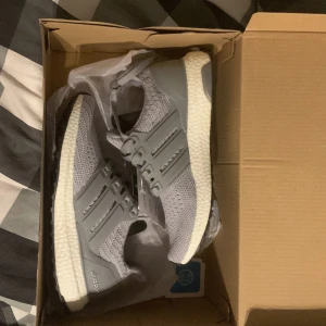 Adidas Ultraboost grå sneakers - Säljer ett par Adidas Ultraboost sneakers i snygg grå färg med stickad ovandel och vita Boost-sulor. Skorna har klassiska tre ränder på sidan, snörning och en rund tå. Perfekta för dig som vill ha både stil och komfort till träning eller häng. Helt oanvända ,säljer pga för dom är små,storlek ca 36/37