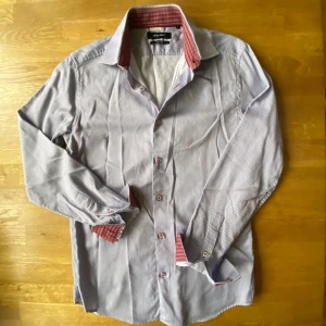 Randig slim fit skjorta Riley Black - Snygg randig skjorta från Riley Black i slim fit-modell. Skjortan är vit med tunna blå och röda ränder, har långa ärmar och klassisk krage med röd-rutigt foder på insidan. Knappar framtill och vid manschetterna. Perfekt för dig som gillar stilrena detaljer.