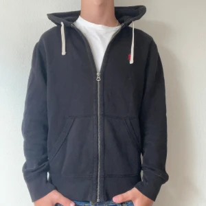 Svart hoodie från Polo Ralph Lauren - Svart hoodie från Polo Ralph Lauren med dragkedja och vit snörning i huvan. Tröjan har en röd broderad logga på bröstet, två fickor fram och är tillverkad i mjuk bomull. Perfekt för chill dagar och enkel att matcha med jeans eller joggers.