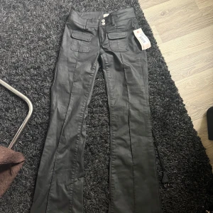 Svarta bootcut byxor från Nelly - Snygga svarta byxor från Nelly med bootcut-snitt och hög midja. Byxorna har två framfickor med lock och dubbla knappar i midjan. Materialet har en lätt glansig finish som ger en cool look. Perfekta för dig som vill sticka ut med stil.