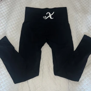 Svarta tränings leggings med hög midja - Svarta träningsbyxor med bred hög midja och ett vitt X-tryck framtill. Stretchigt material som sitter tight och är perfekt för träning. Snygg och enkel design som funkar till allt, med mjuk och följsam känsla. Formar bra! 😊